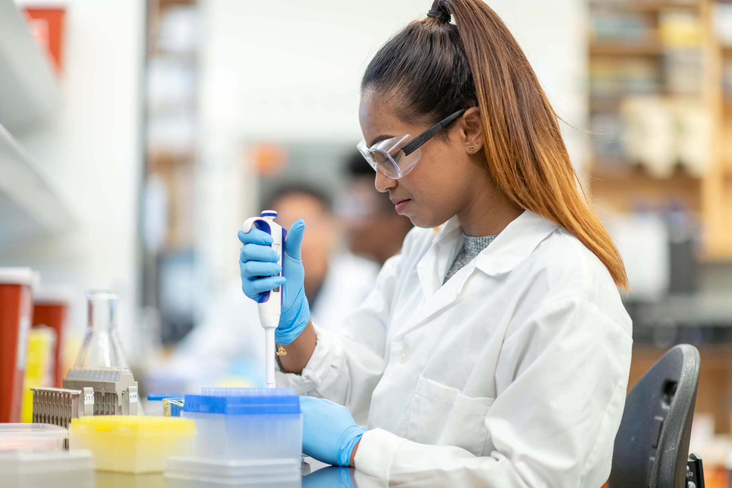 A scientist in safety glasses using a pipette at a laboratory bench.