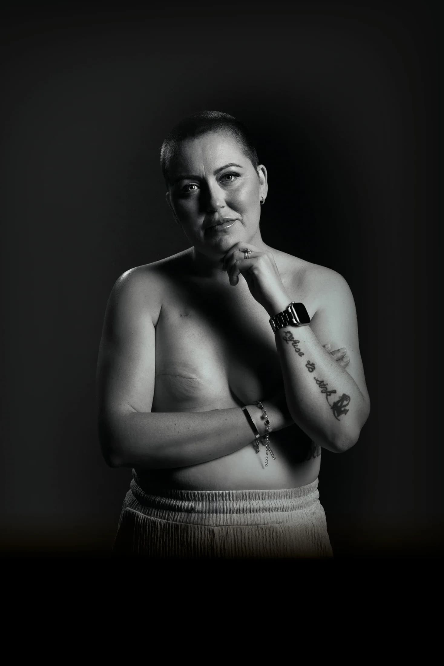 A black and white Breast Cancer Know More campaign portrait of a woman with a shaved head, her hand at her chin.